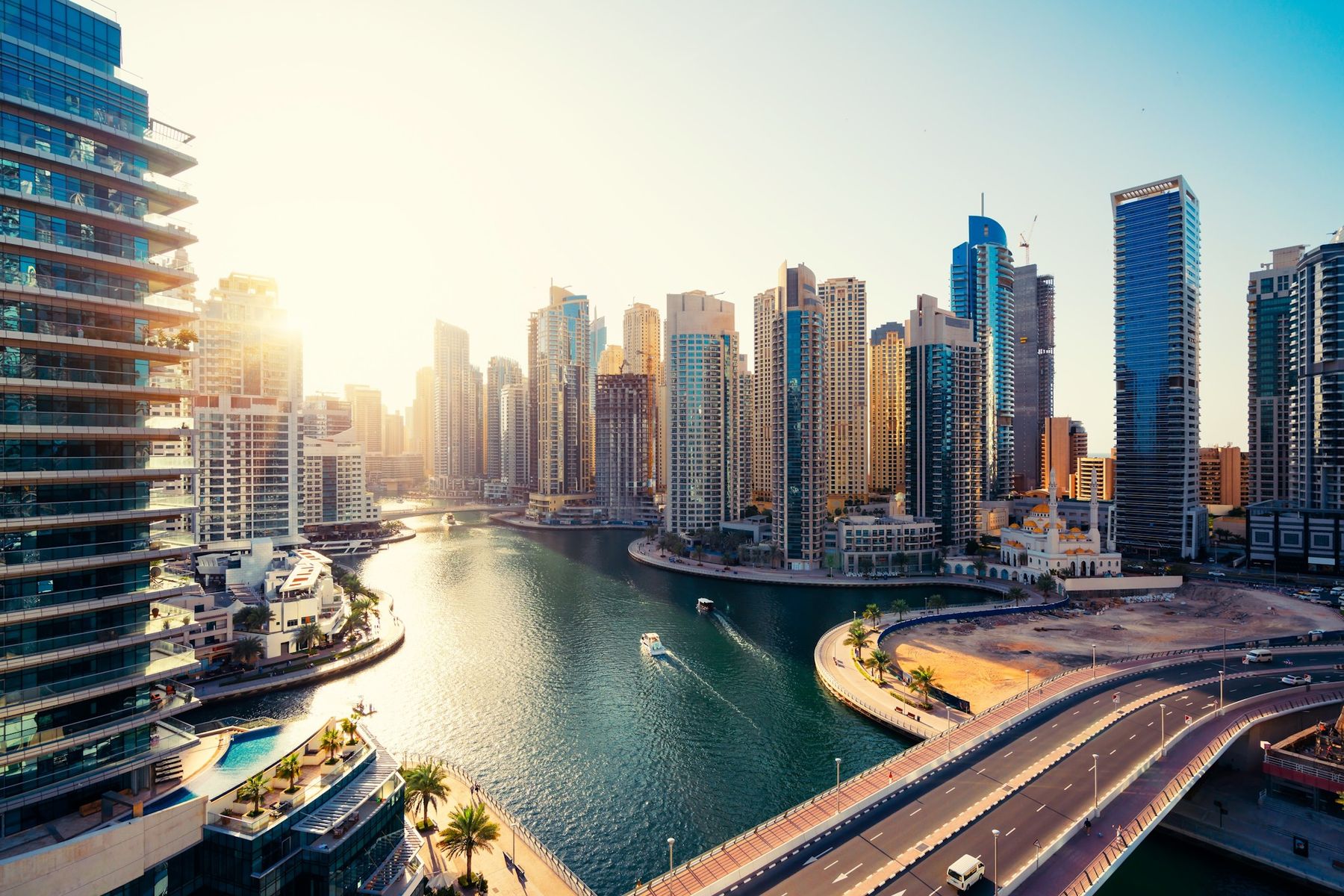 Glass and sandstone residential towers lining a curving marina waterway, with a bridge and small boats in the foreground at sunset