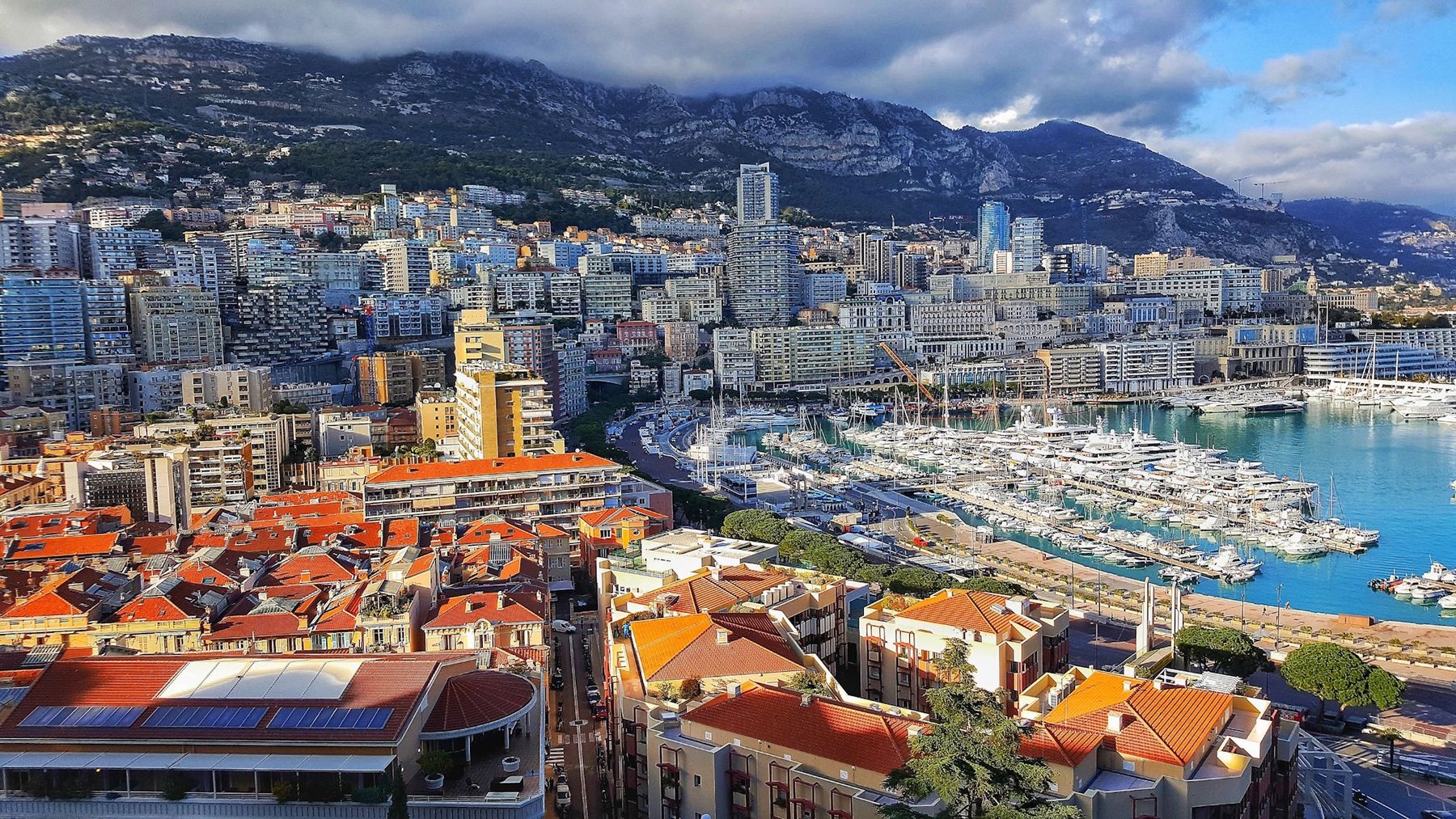 Aerial view of a Mediterranean harbour lined with white yachts, with apartment blocks and mountains behind